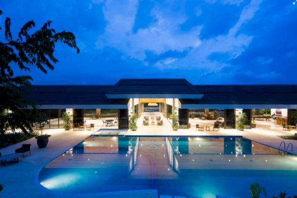 A luxurious, modern building with bright outdoor lighting overlooks a large pool under a twilight sky.