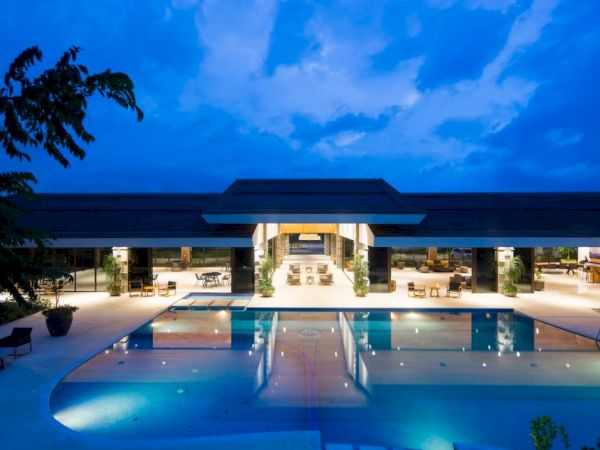 A luxurious, modern building with bright outdoor lighting overlooks a large pool under a twilight sky.