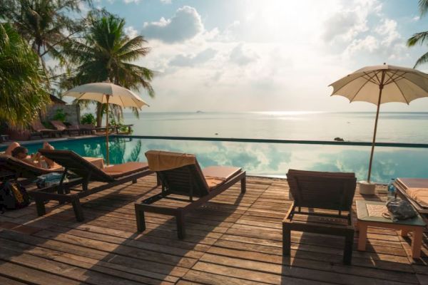 Lounge chairs and umbrellas by a pool overlook a calm sea, with palm trees and a cloudy sky creating a relaxing tropical scene.