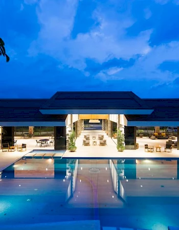 A luxurious building with large outdoor pool and lounge area, illuminated at night under a cloudy blue sky.