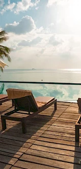 A tranquil scene with lounge chairs by a pool, umbrellas, palm trees, and a view of the ocean under a partly cloudy sky.