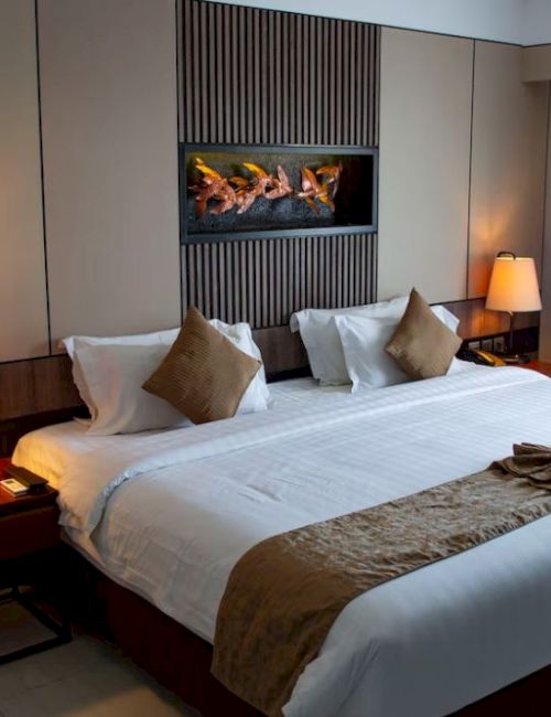 A neatly arranged hotel room features a large bed with decorative pillows, lamps on side tables, and a wall-mounted fireplace.