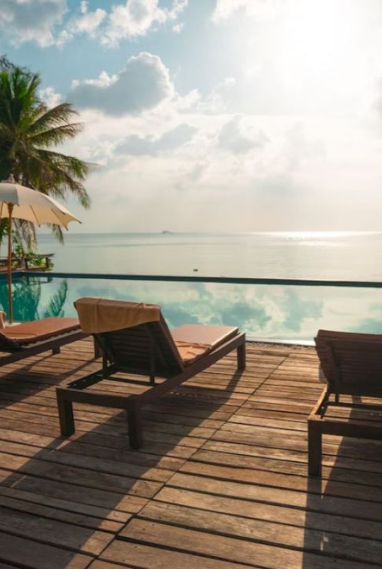 Sunlit deck with loungers and umbrellas overlooks a serene infinity pool and ocean, set against a backdrop of palm trees and cloudy sky.