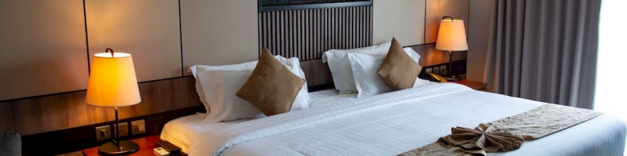 A well-decorated hotel room features a large bed with cushions, a wall-mounted art piece, lamps, and curtains for a cozy ambiance.