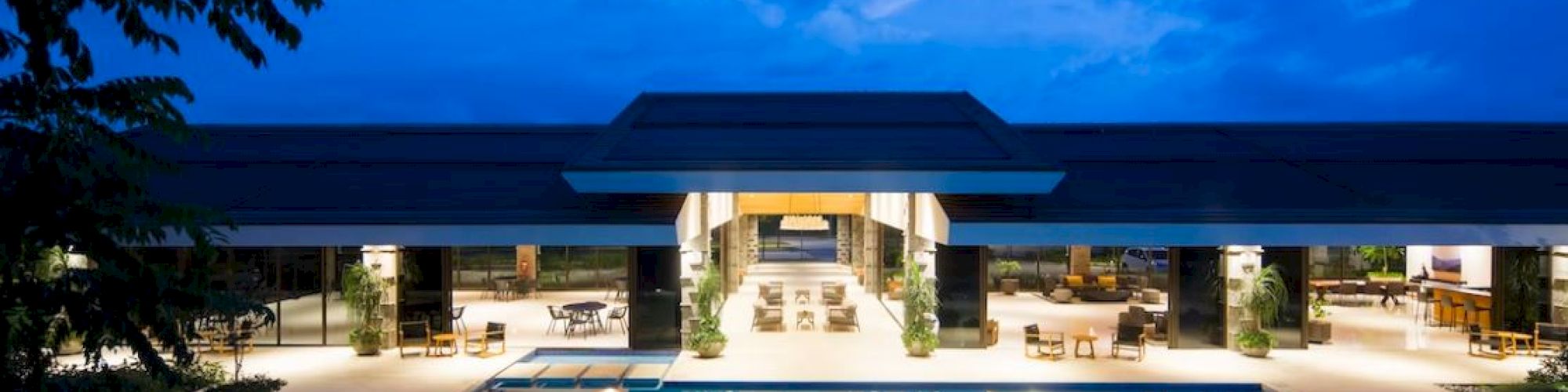 The image shows a luxurious modern building with an inviting swimming pool, illuminated under a dusky blue sky.