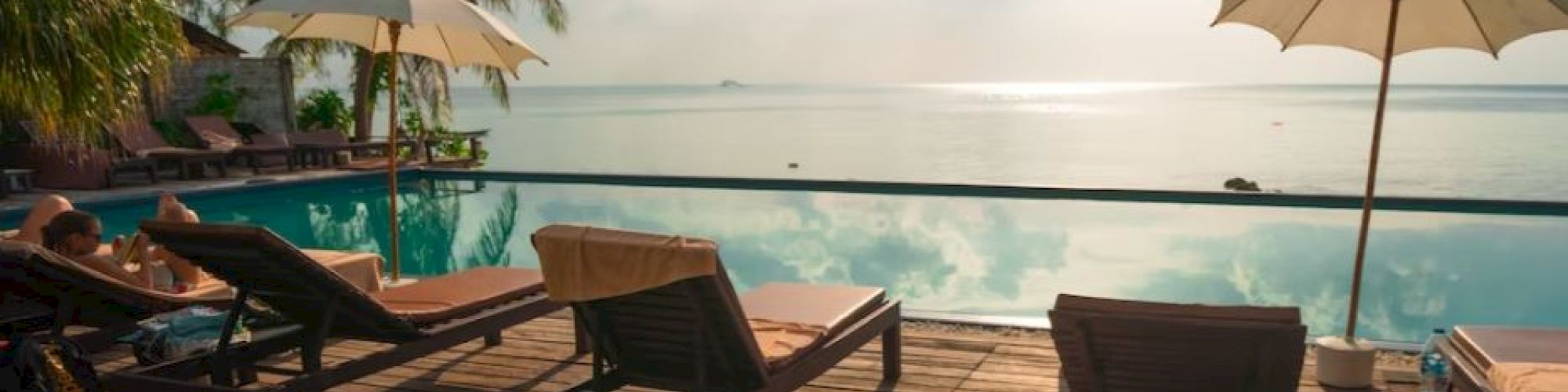 A tropical poolside scene with lounge chairs, umbrellas, palm trees, and a view of the ocean under a bright sky with scattered clouds.