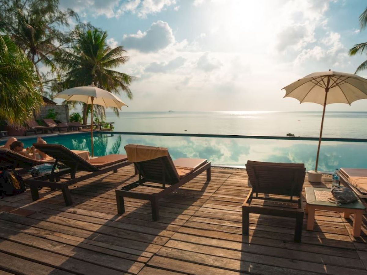 A serene poolside scene with lounge chairs, umbrellas, and palm trees overlooking calm water under a bright sky.