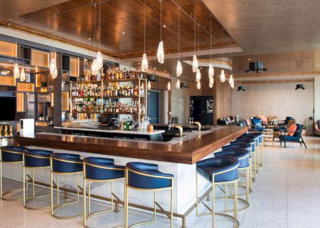 A modern bar with a central counter, stylish bar stools, hanging lights, and a well-stocked liquor shelf in a chic interior setting.