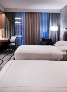 The image shows a modern hotel room with two beds, a wall-mounted TV, a desk with a chair, and stylish lighting.
