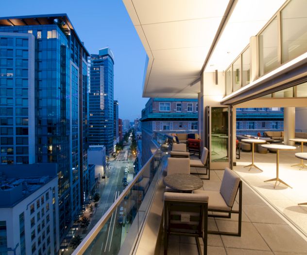 A modern rooftop patio at dusk with lounge seating and tables, overlooking a city street lined with tall buildings.
