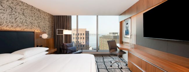 A modern hotel room with a large bed, TV, desk, and chair, featuring a cityscape view from the window.