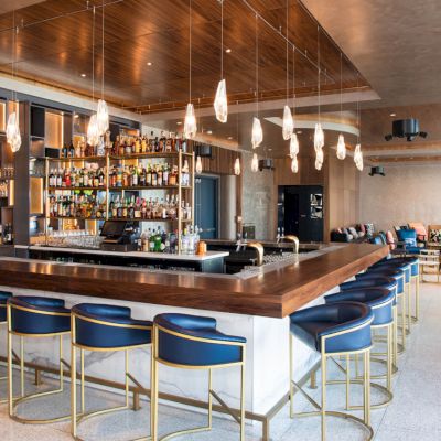 The image shows a modern bar with a central counter, high chairs, hanging lights, and a stocked liquor shelf in a stylish lounge setting.