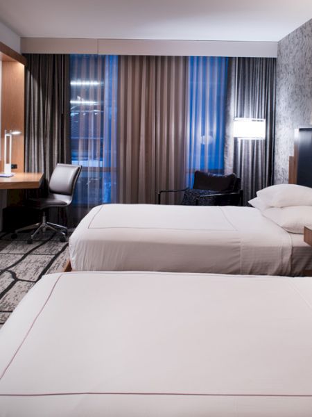 The image shows a modern hotel room with two beds, a wall-mounted TV, desk, chair, and stylish decor, featuring patterned wallpaper and carpet.