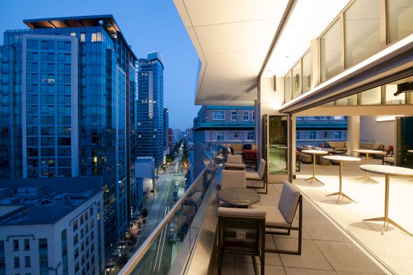The image shows a modern balcony with seating, overlooking a cityscape with tall buildings, during evening hours.