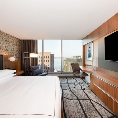 A modern hotel room features a large bed, TV, desk, and chair with a city view through the window, offering a sleek and comfortable design.