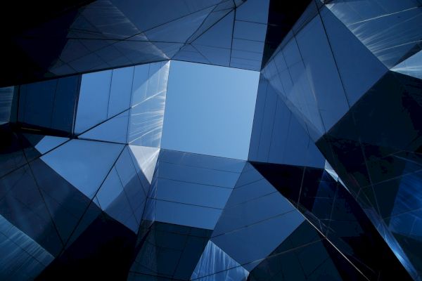 The image shows a geometric, abstract view of skyscrapers with glass and metal surfaces reflecting the sky, forming a complex structure.