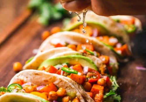 A hand squeezes lime over chickpea and vegetable tacos, garnished with cilantro and avocado slices, with lime wedges on a wooden board.