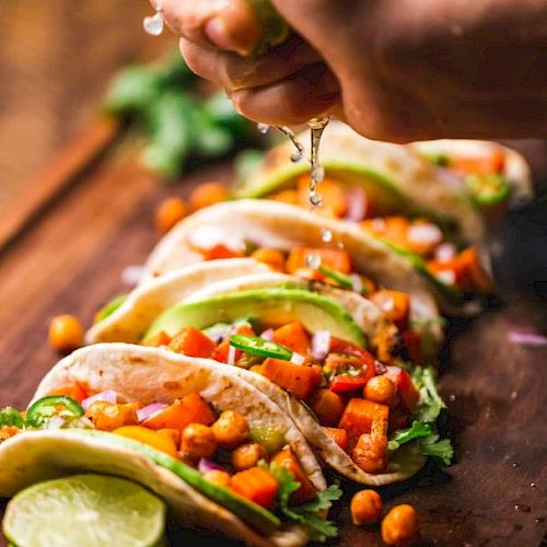 A hand squeezes lime over chickpea and vegetable tacos, garnished with cilantro and avocado slices, with lime wedges on a wooden board.
