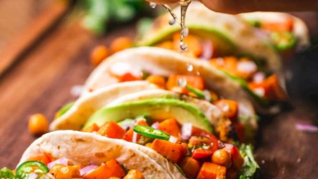 A hand squeezes lime over chickpea and vegetable tacos, garnished with cilantro and avocado slices, with lime wedges on a wooden board.