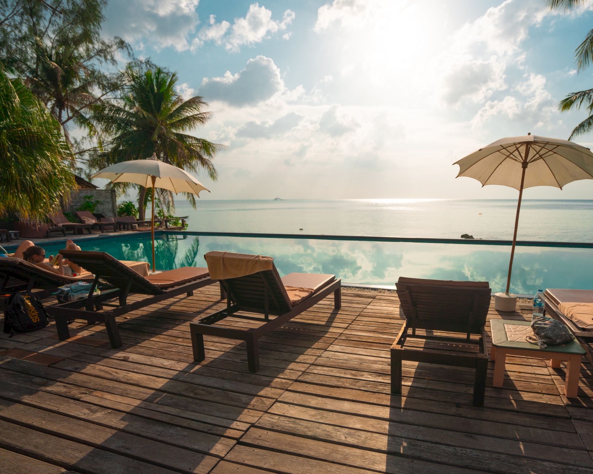 A tranquil beach scene with lounge chairs, umbrellas, an infinity pool, and a view of the ocean under a partly cloudy sky.