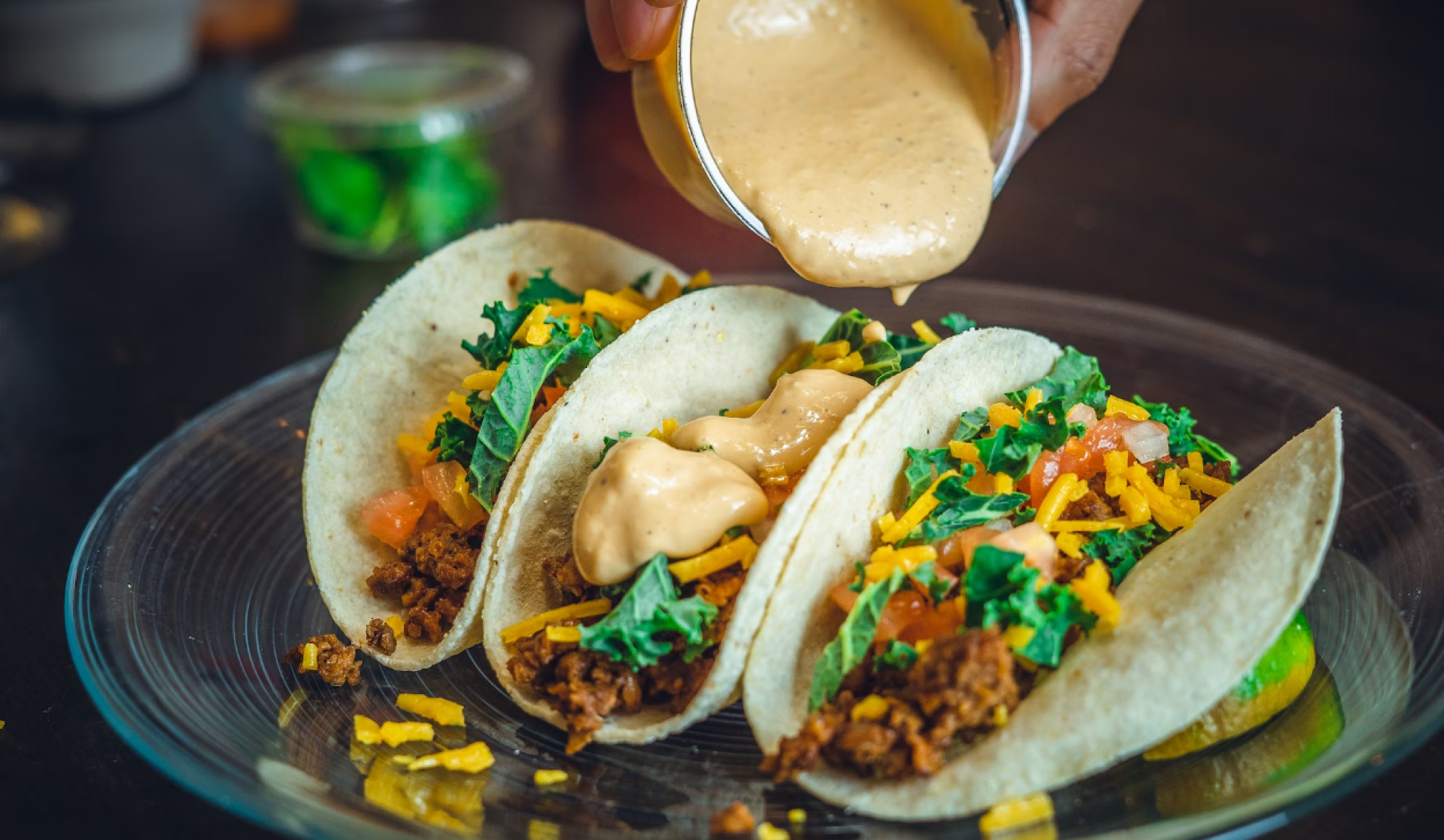 Three tacos with ground meat, cheese, lettuce, and salsa are on a plate, with sauce being poured over them.