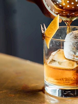 A drink being poured into a glass with ice and a citrus peel garnish, likely a cocktail, on a bar counter.