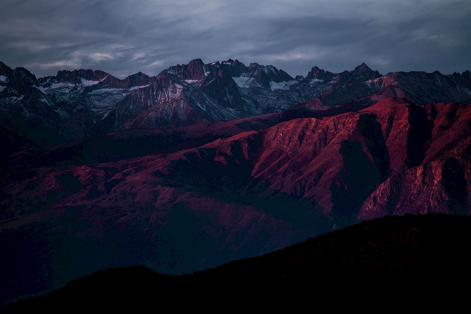 A mountain range at sunset with dramatic lighting casting a reddish hue over the peaks and valleys under a cloudy sky.
