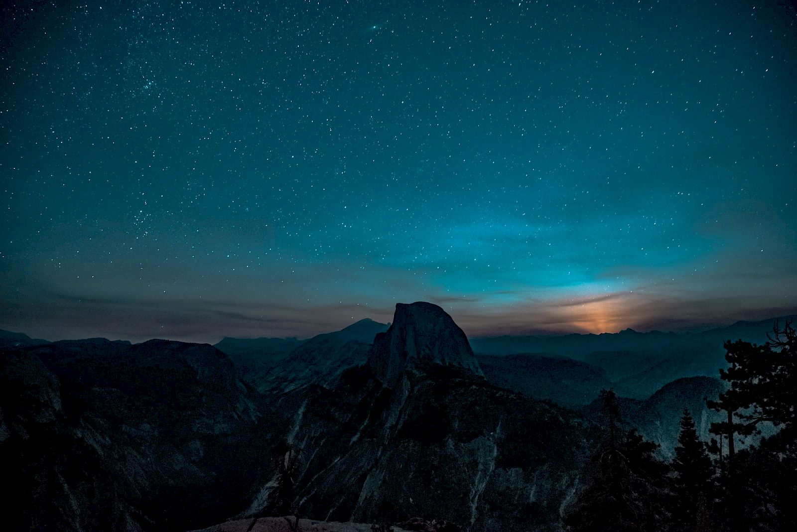 A mountainous landscape under a starry night sky with a faint glow, possibly moonrise or sunrise, casting light over the peaks.