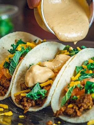 Three tacos filled with ground meat, shredded cheese, and vegetables being topped with a creamy sauce.
