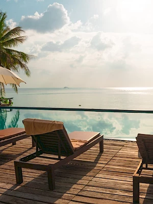 A serene beachside pool scene with lounge chairs, umbrellas, and palm trees overlooking a calm ocean under a partly cloudy sky.