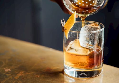 A bartender pours a brown cocktail into a glass with ice and a garnish.