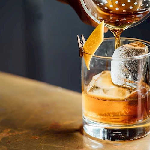 A bartender pours a brown cocktail into a glass with ice and a garnish.