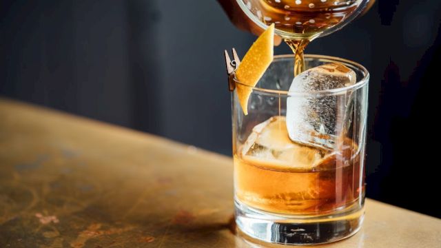 A bartender pours a brown cocktail into a glass with ice and a garnish.