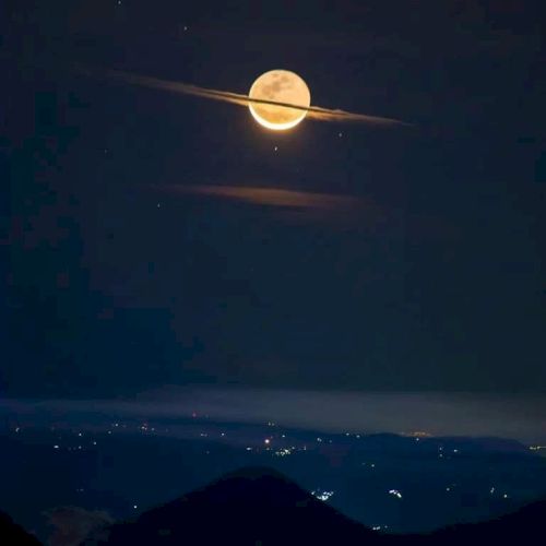 A full moon with clouds resembling Saturn's rings shines over a landscape with scattered lights, creating a celestial illusion.