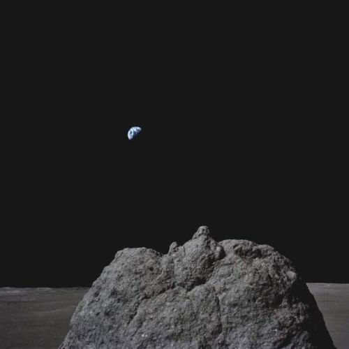 A large rock on the Moon's surface with Earth visible in the distant black sky above it.