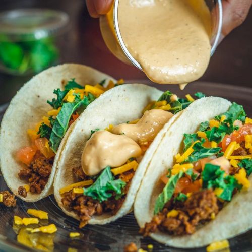 Three tacos with ground meat, cheese, lettuce, and tomatoes are being topped with sauce on a plate.