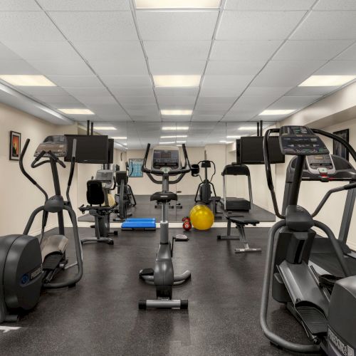 A gym with exercise machines, such as ellipticals, stationary bikes, weights, and fitness balls, with mirrors and TVs on walls.