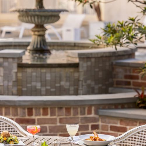 Outdoor dining setup with dishes, cocktails on a wooden table, near a fountain, and leafy surroundings.
