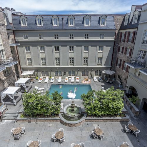 A courtyard with a pool surrounded by lounge chairs, cabanas, and greenery, enclosed by multi-story buildings.
