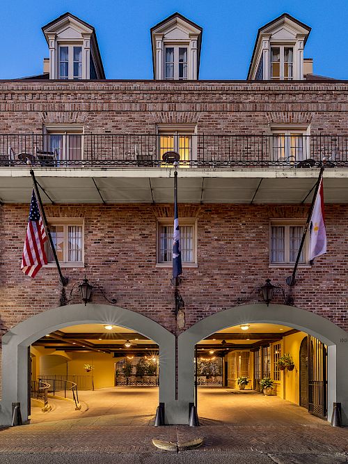 A building with balconies, arched entrance, and hanging flags is shown. It features traditional architecture with decorative railings.