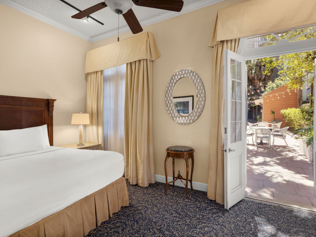 A bright bedroom with a bed, beige curtains, ceiling fan, round wall mirror, and an open door to a sunny patio with chairs.