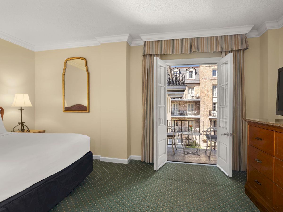 A hotel bedroom with a bed, dresser, wall mirror, and open French doors with curtains leading to a balcony overlooking brick buildings.