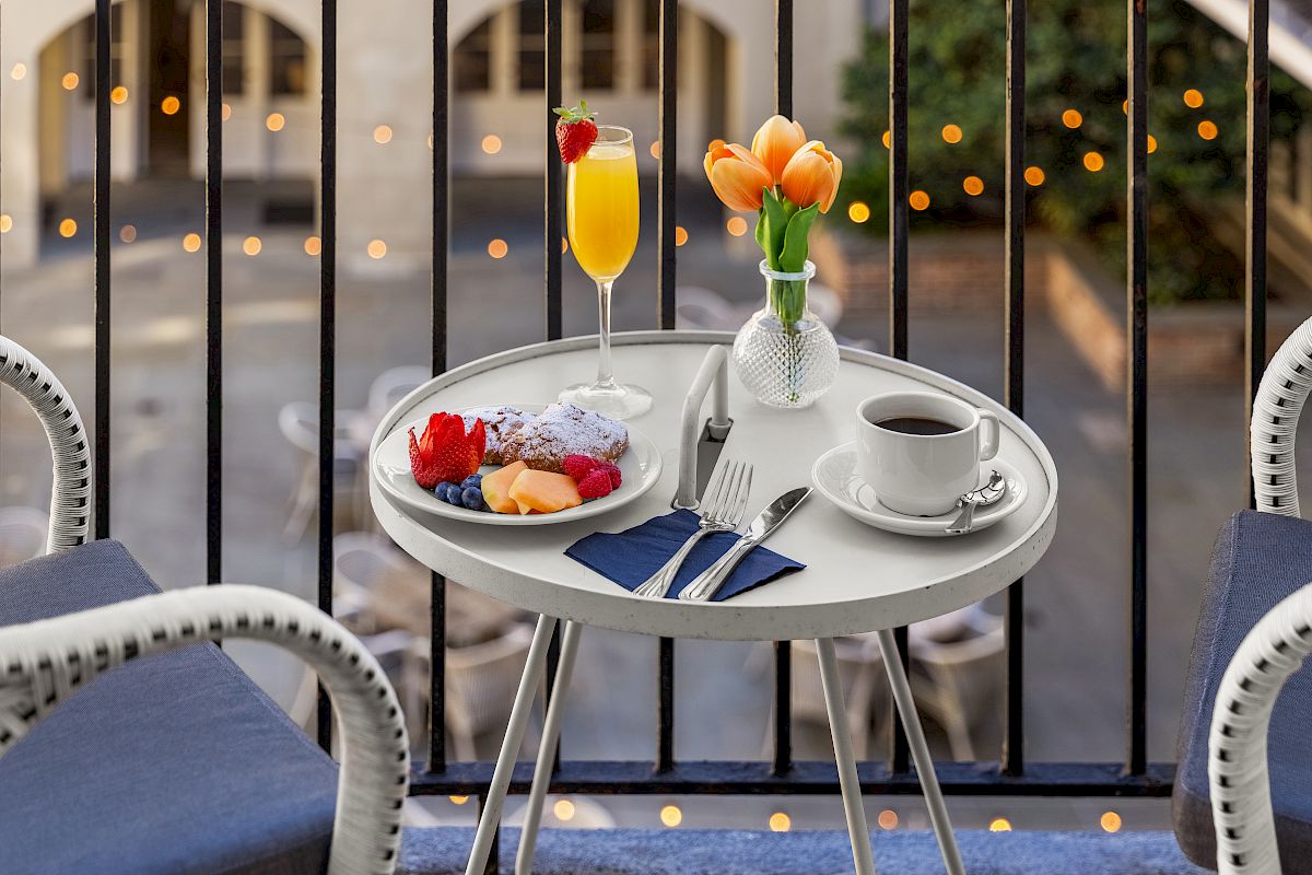 A small round table with breakfast items, including fruit, coffee, and a drink, on a balcony with a view of a courtyard, decorated with tulips.