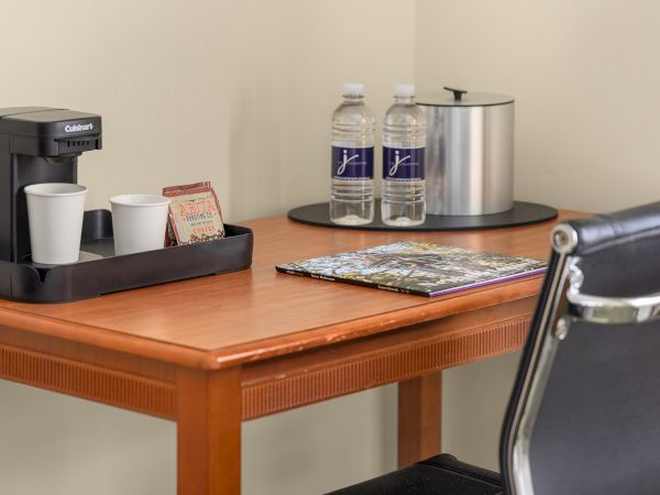 A coffee maker with cups, coffee packets, two water bottles, an ice bucket, and a magazine on a wooden desk.