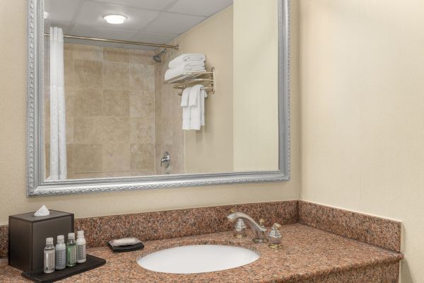 A hotel bathroom featuring a sink, toiletries, a tissue box, a mirror, and a shower area with folded towels on a rack.