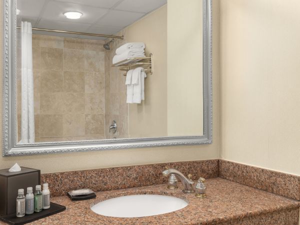 A hotel bathroom featuring a sink, toiletries, a tissue box, a mirror, and a shower area with folded towels on a rack.