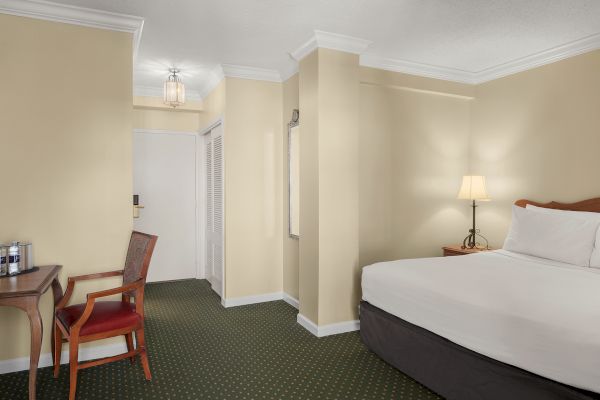 A hotel room with a bed, side table, lamp, chair, desk, mirror, and white door, set against beige walls and green carpet.