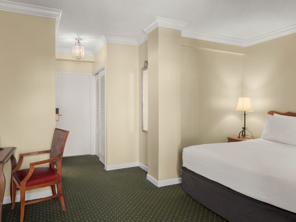 A hotel room with a bed, side table, lamp, chair, desk, mirror, and white door, set against beige walls and green carpet.