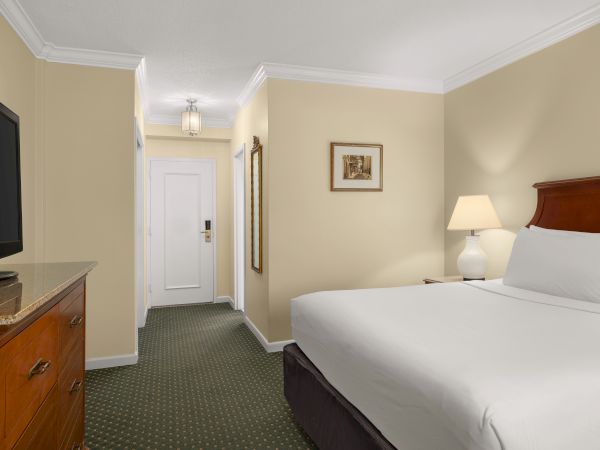 A hotel room with a bed, TV, and lamp. There's a framed picture on the wall near the entrance and a light fixture above.