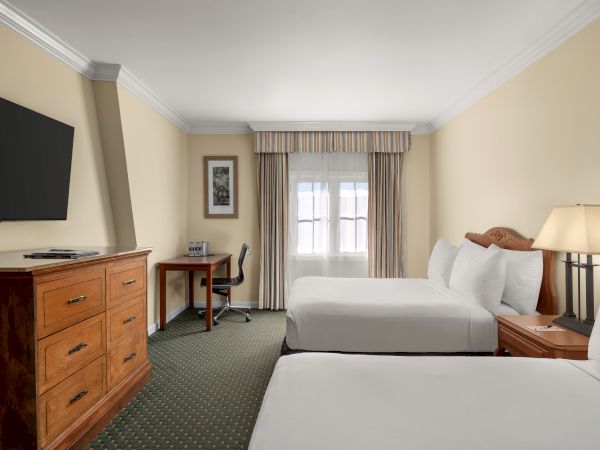 A hotel room with two beds, a TV on a dresser, a desk with a chair, and a lamp by the window with curtains.